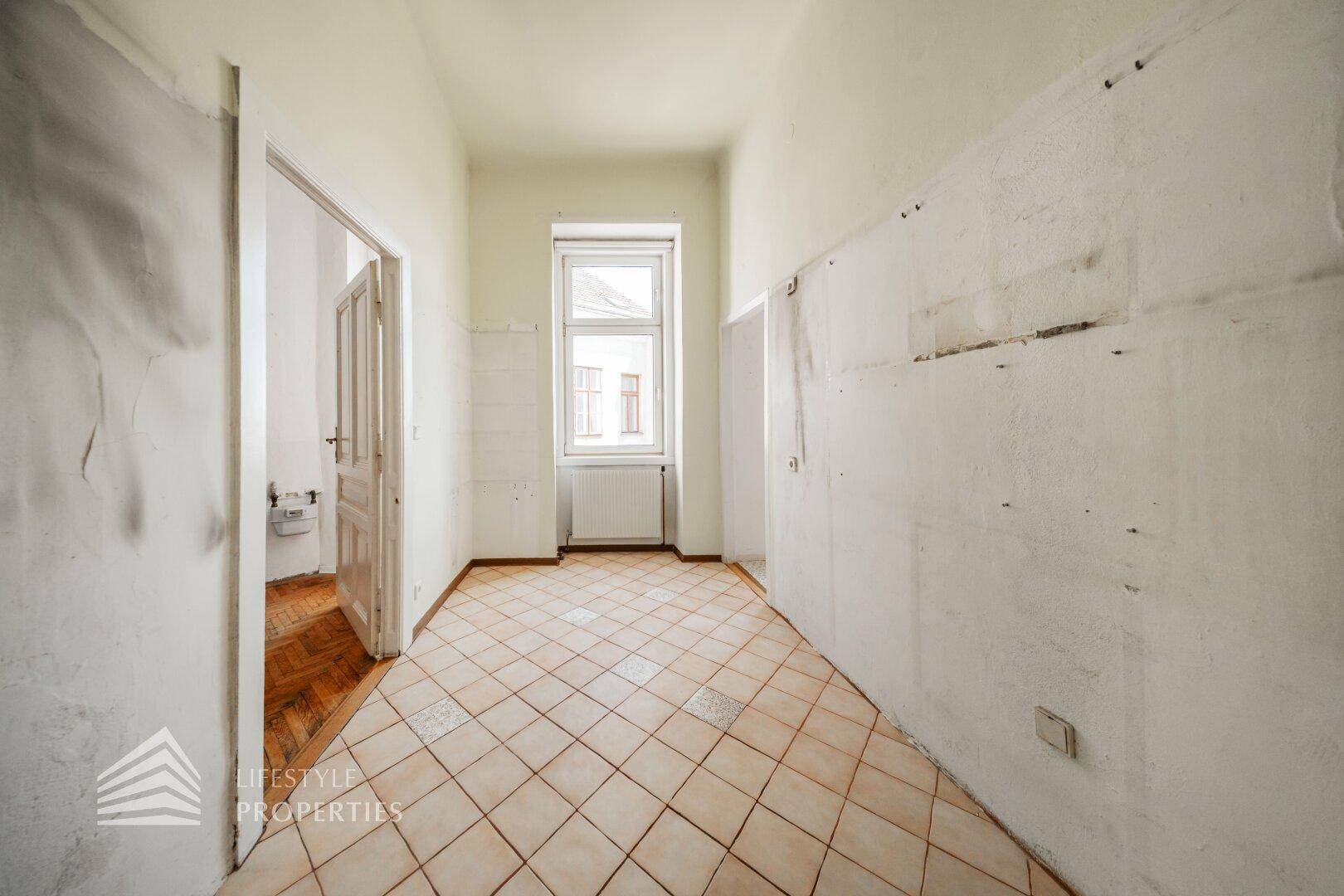 Helle, leere Küche mit Fenster, alten Fliesen und Spuren einer ehemaligen Küchenzeile.