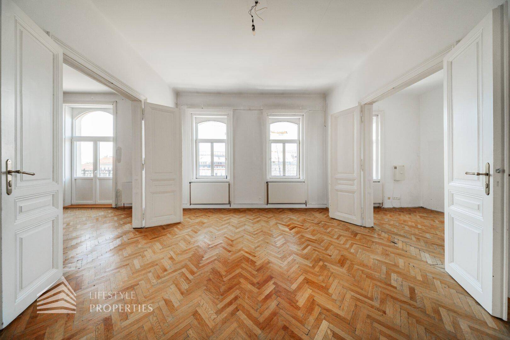Großzügiges Wohnzimmer mit Fischgrätparkett, hohen Fenstern und Flügeltüren, viel Tageslicht.