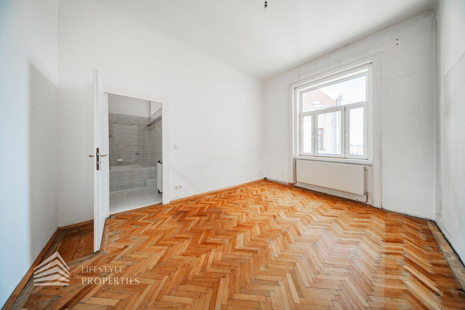 Helles Schlafzimmer mit Fischgrätparkett und Fenster, angrenzend an ein Badezimmer.
