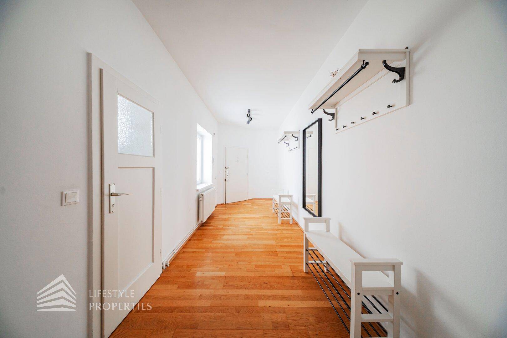 Langer Flur mit Holzboden, weißer Garderobe und einem Fenster, das Tageslicht spendet.