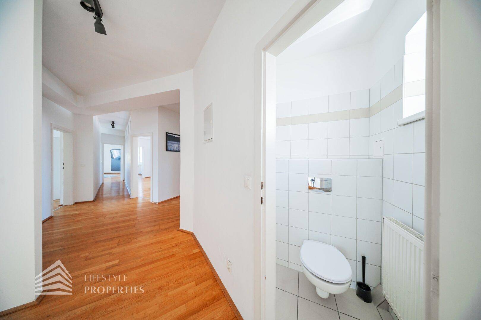 Separate Toilette mit weißen Fliesen, wandhängendem WC und einem kleinen Fenster für Belüftung.
