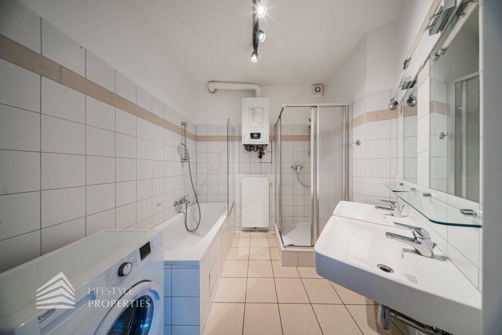 Badezimmer mit weißen Fliesen, Badewanne, Dusche, Doppelwaschbecken und Waschmaschine.