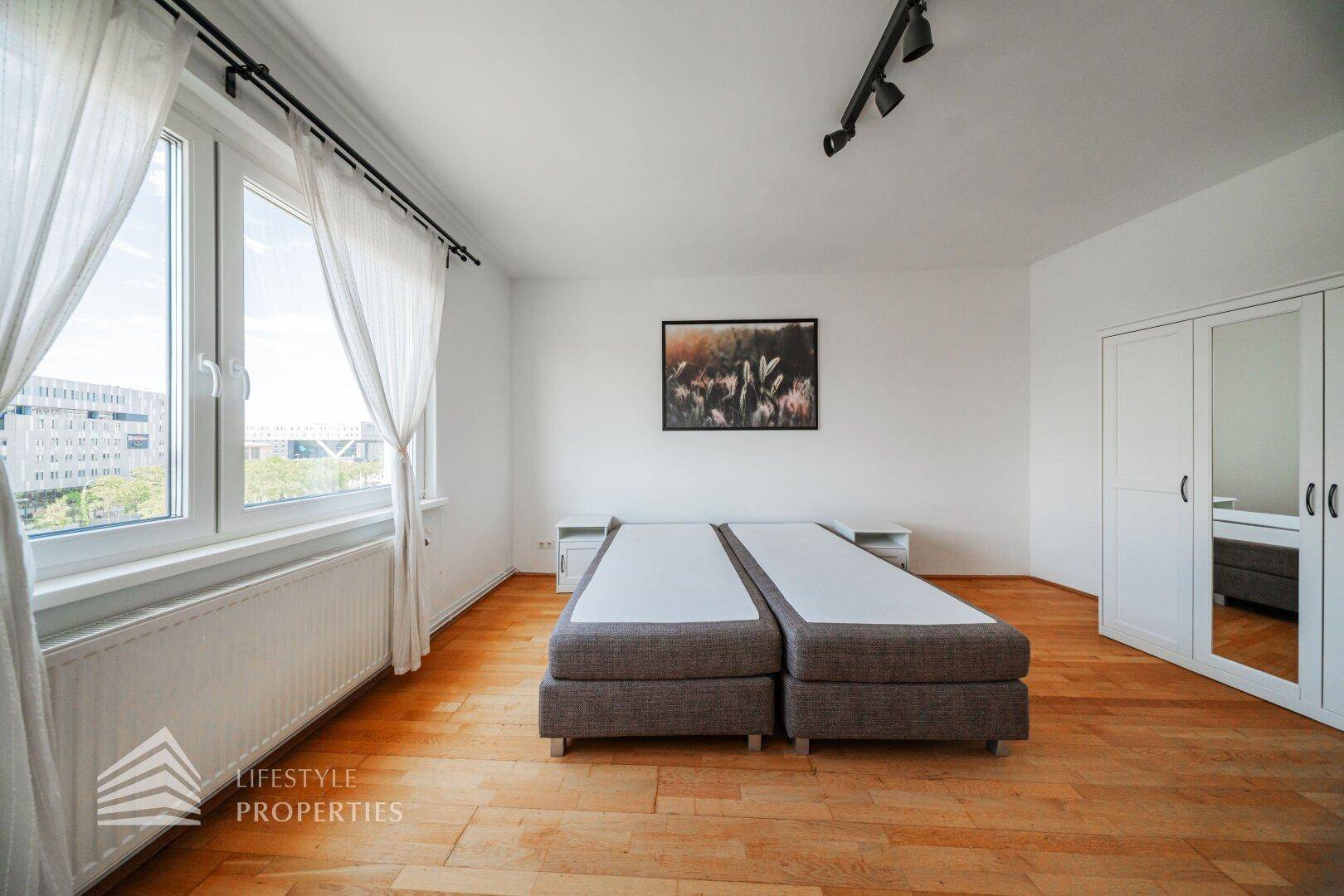 Schlafzimmer mit großen Fenstern, Holzboden und zwei Einzelbetten, die viel Tageslicht hereinlassen.