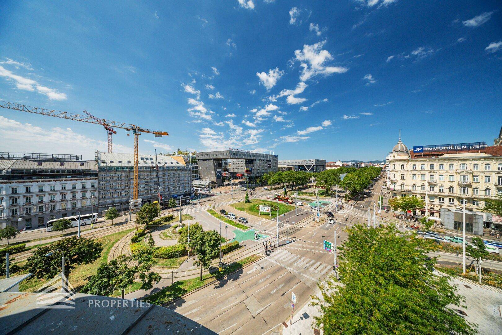 Weitläufiger Stadtblick über eine belebte Kreuzung mit Gebäuden und viel Grün.