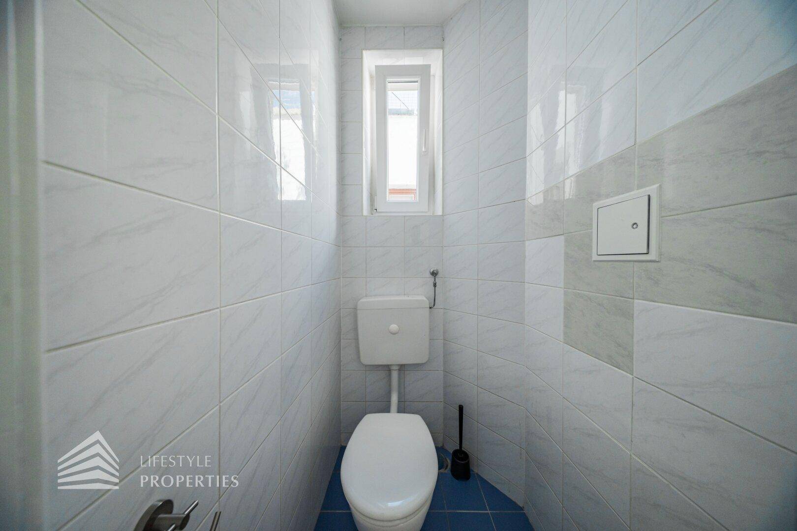 Kleines WC mit weißen Fliesen, Toilette und einem schmalen Fenster für Tageslicht.