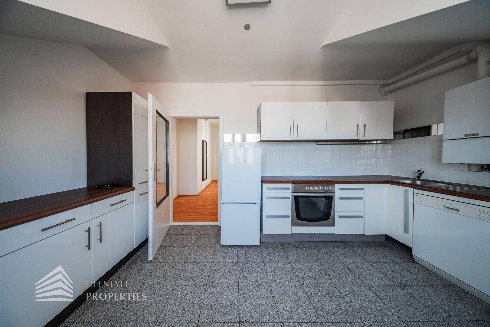 Geräumige Küche mit weißen Schränken, Backofen und Kühlschrank, offen zum Flur.