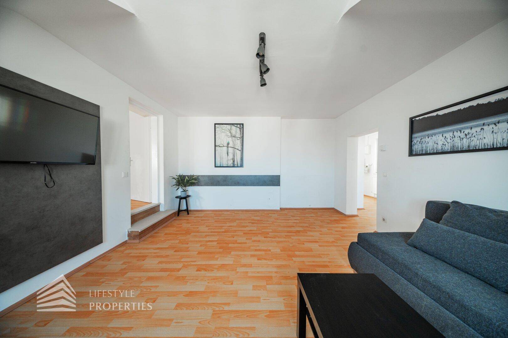 Geräumiges Wohnzimmer mit Holzfußboden, Fernseher und Blick in angrenzende Räume.