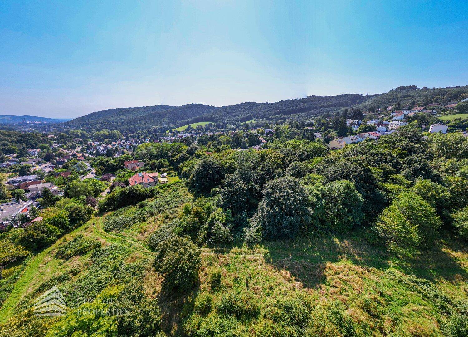 Luftaufnahme einer Wohnsiedlung umgeben von üppiger grüner Natur und sanften Hügeln unter blauem Himmel.