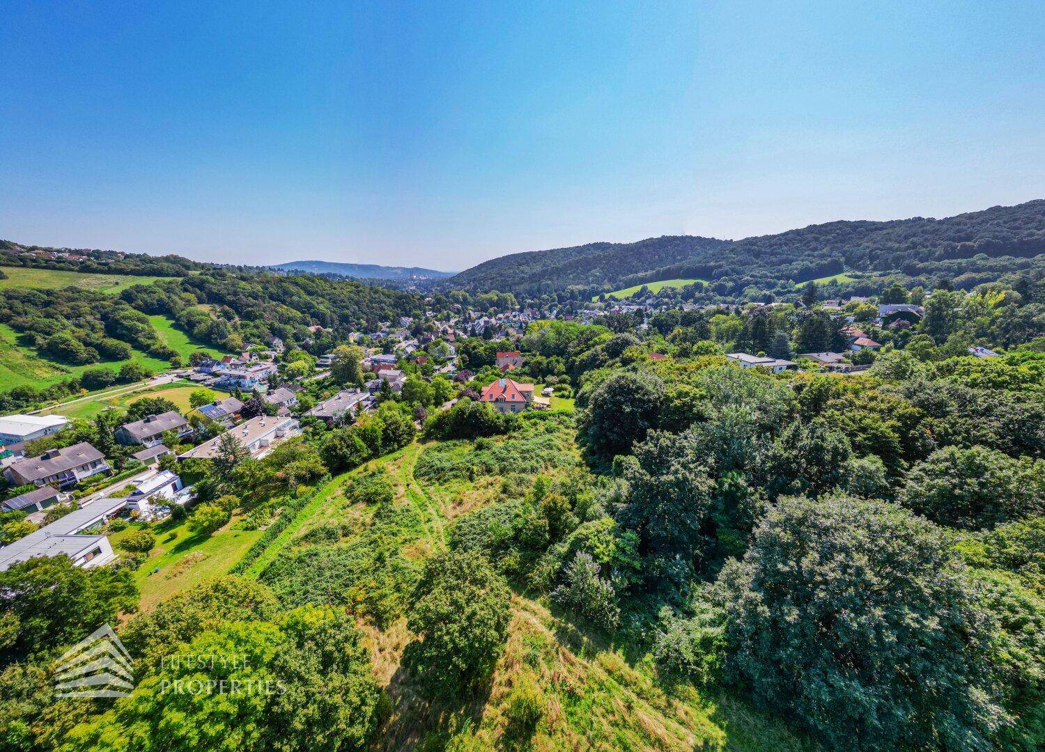 Weitläufige Luftaufnahme einer Siedlung eingebettet in grüne Hügel und Wälder unter blauem Himmel.