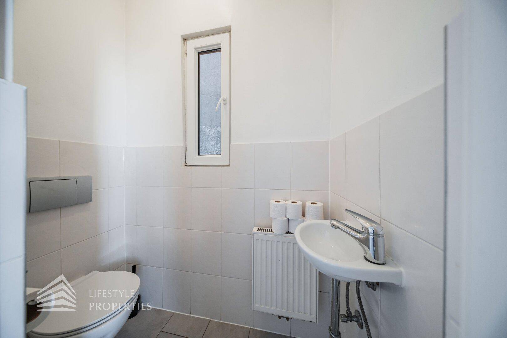 Kleines Gäste-WC mit Toilette, Handwaschbecken und Fenster, hell gefliest.