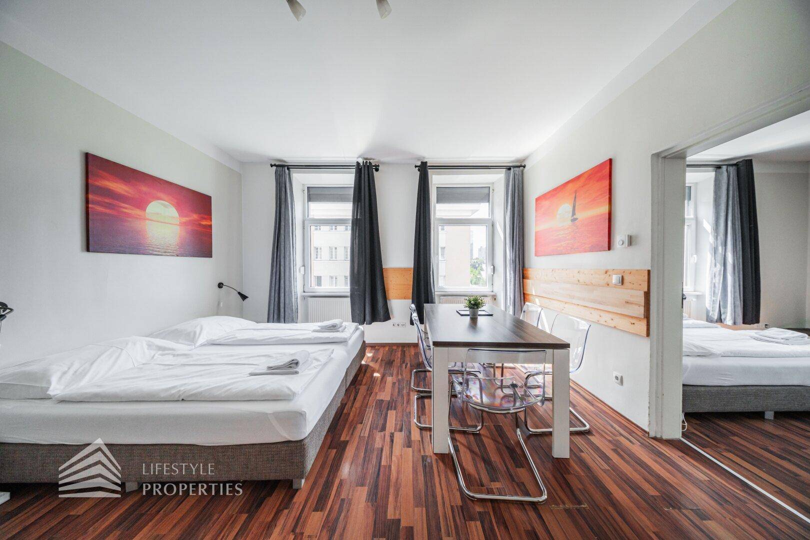 Geräumiges Studio-Apartment mit Holzboden, Doppelbett und Esstisch, durch große Fenster gut belichtet.