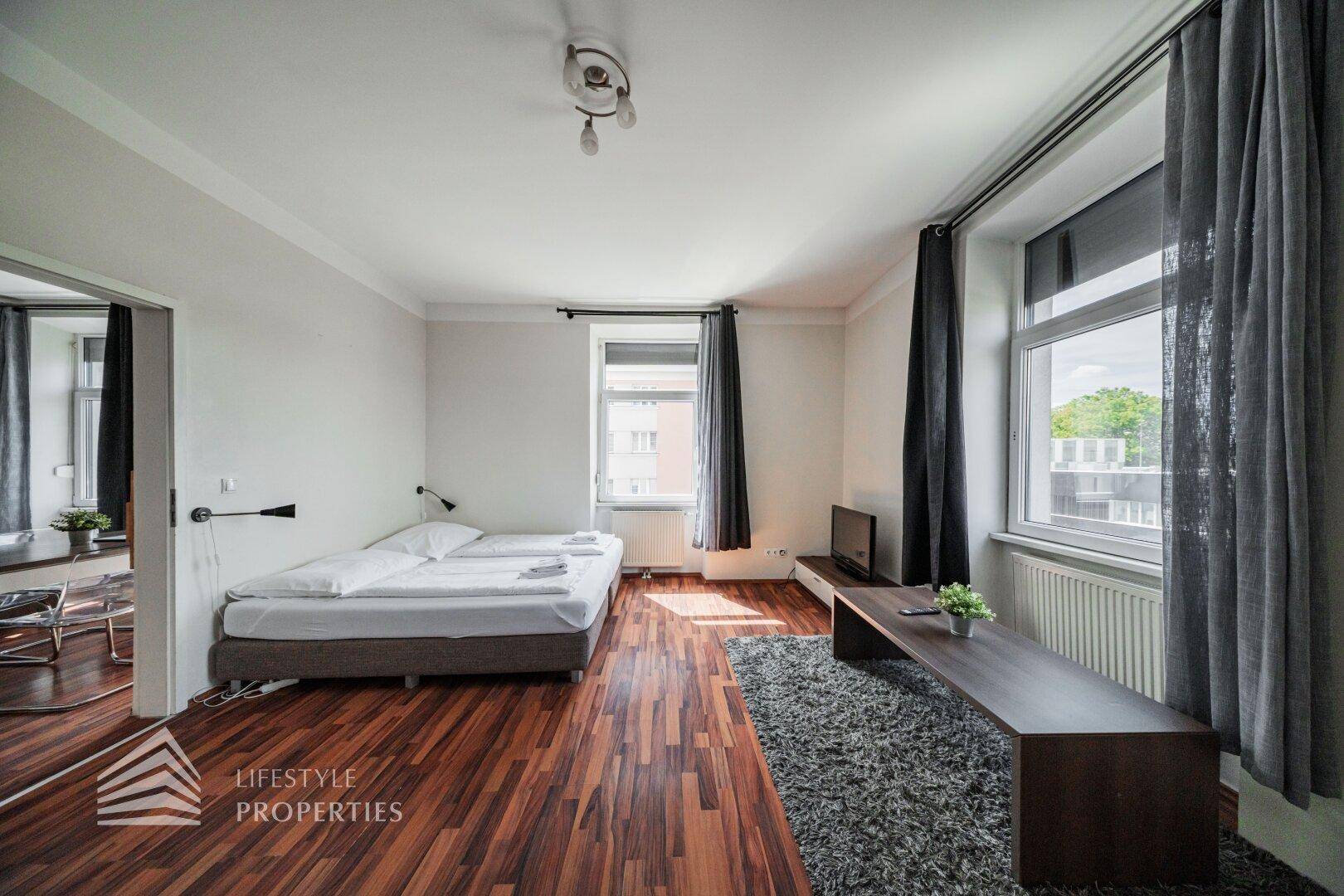 Gemütlicher Wohn- und Schlafbereich mit Holzboden, Doppelbett und Blick aus den großen Fenstern.