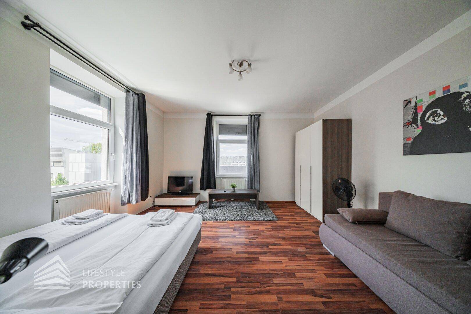 Heller Wohnbereich mit Schlafsofa, Doppelbett und Kleiderschrank, durch große Fenster gut belichtet.