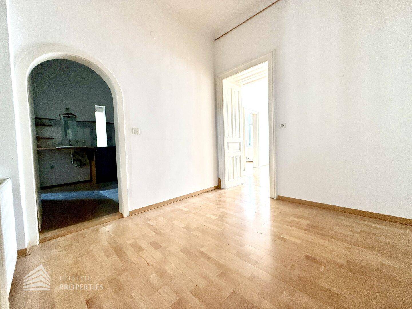 Geräumiges Zimmer mit Holzdielenboden und Rundbogen, führt zu einem weiteren Raum mit Spülbecken.
