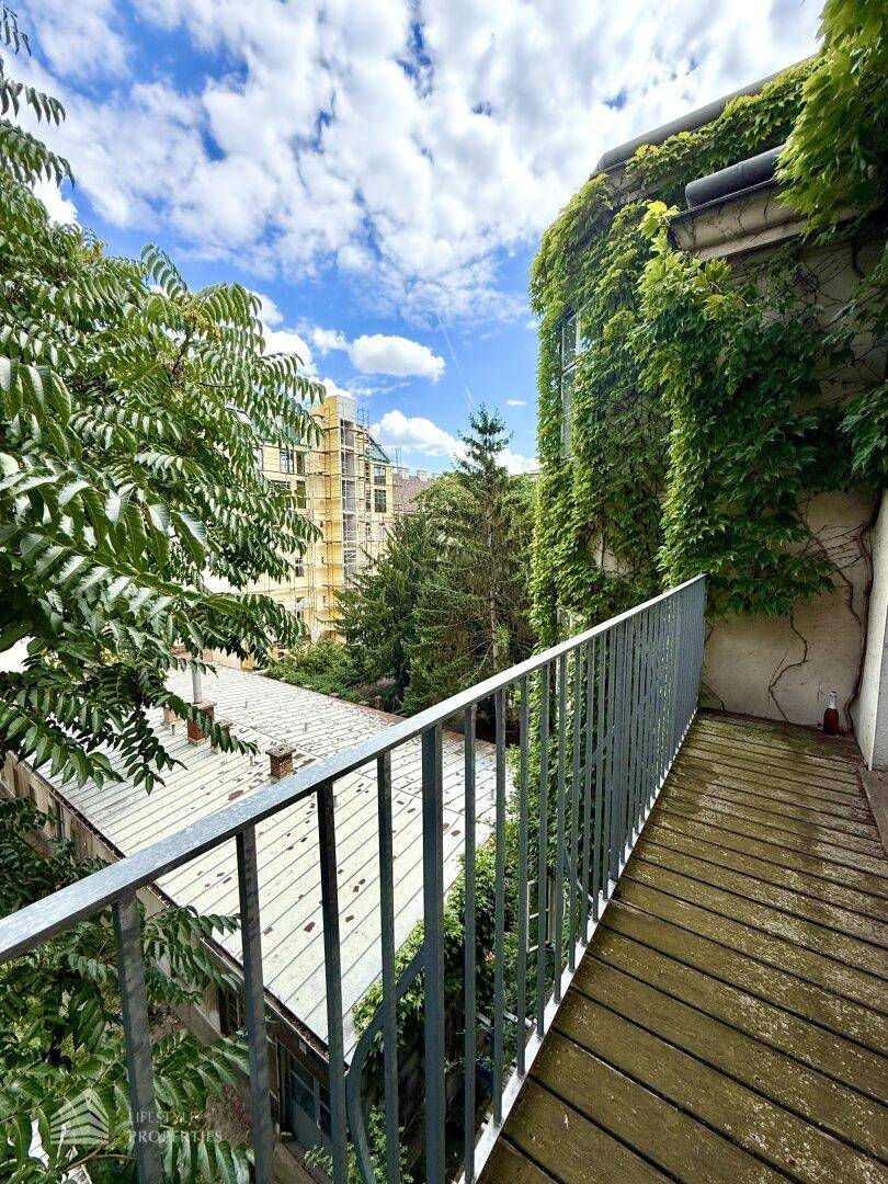 Langer Balkon mit Holzdielen und Blick auf Bäume sowie umliegende Gebäude.
