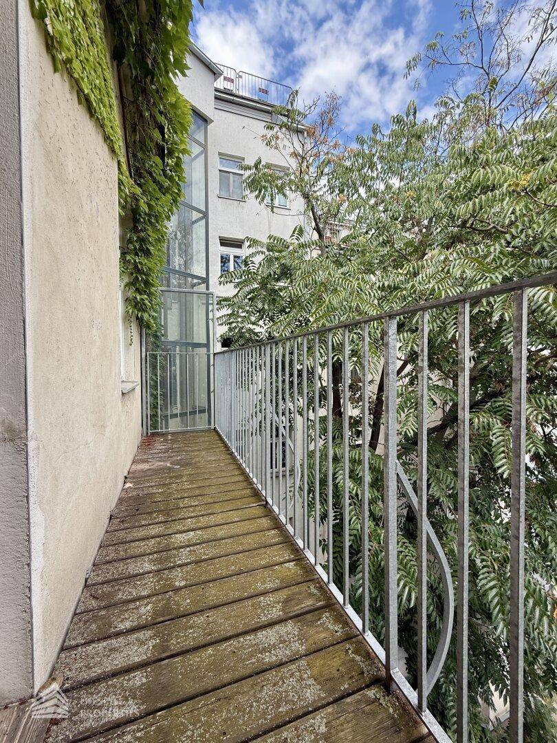 Schmaler Balkon mit Holzboden und Metallgeländer, umgeben von üppigem Grün und einem Glasaufzug.