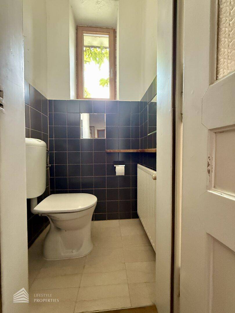 Funktionelles WC mit Toilette, Heizung und Fenster, gestaltet mit dunklen und hellen Wandfliesen.
