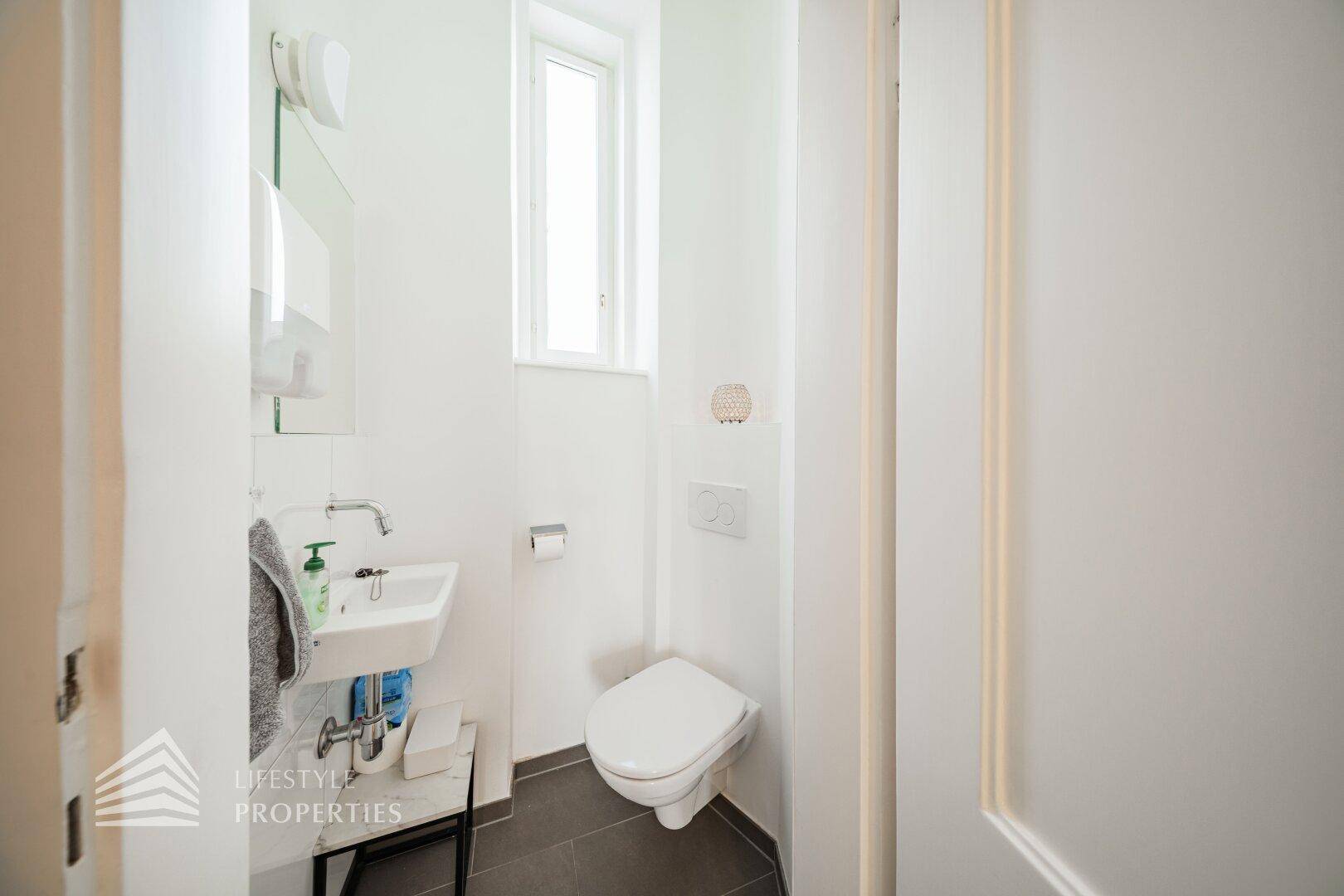 Kleines Gäste-WC mit wandhängender Toilette, Handwaschbecken und einem Fenster für natürliche Belüftung.