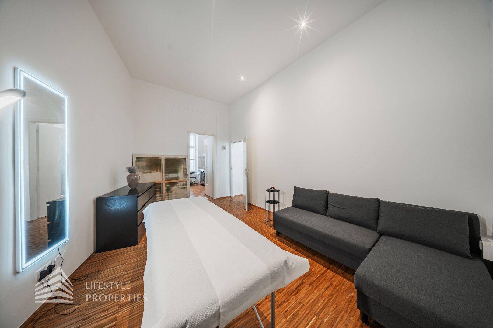 Moderner Behandlungsraum mit grauem Sofa, Behandlungsliege und einem großen, beleuchteten Spiegel an der Wand.