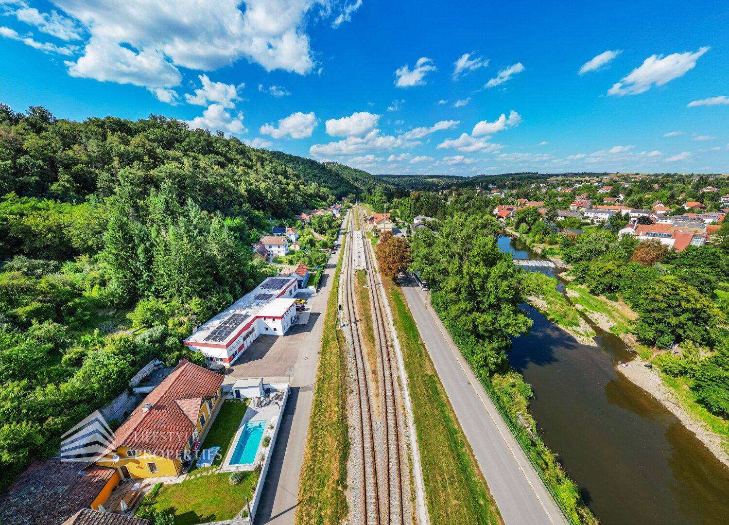 Luftaufnahme einer ländlichen Gegend mit Gewerbegebäude, Bahnlinie, Fluss und umliegenden Häusern.