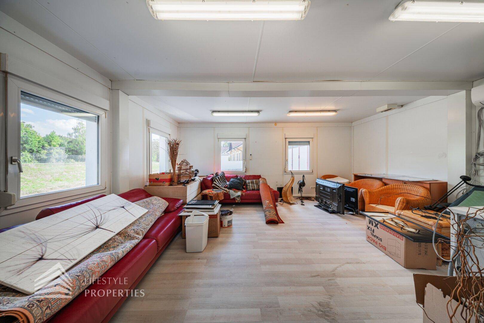 Geräumiger Büroraum mit hellem Laminatboden und großen Fenstern, teilweise möbliert mit Sofas.