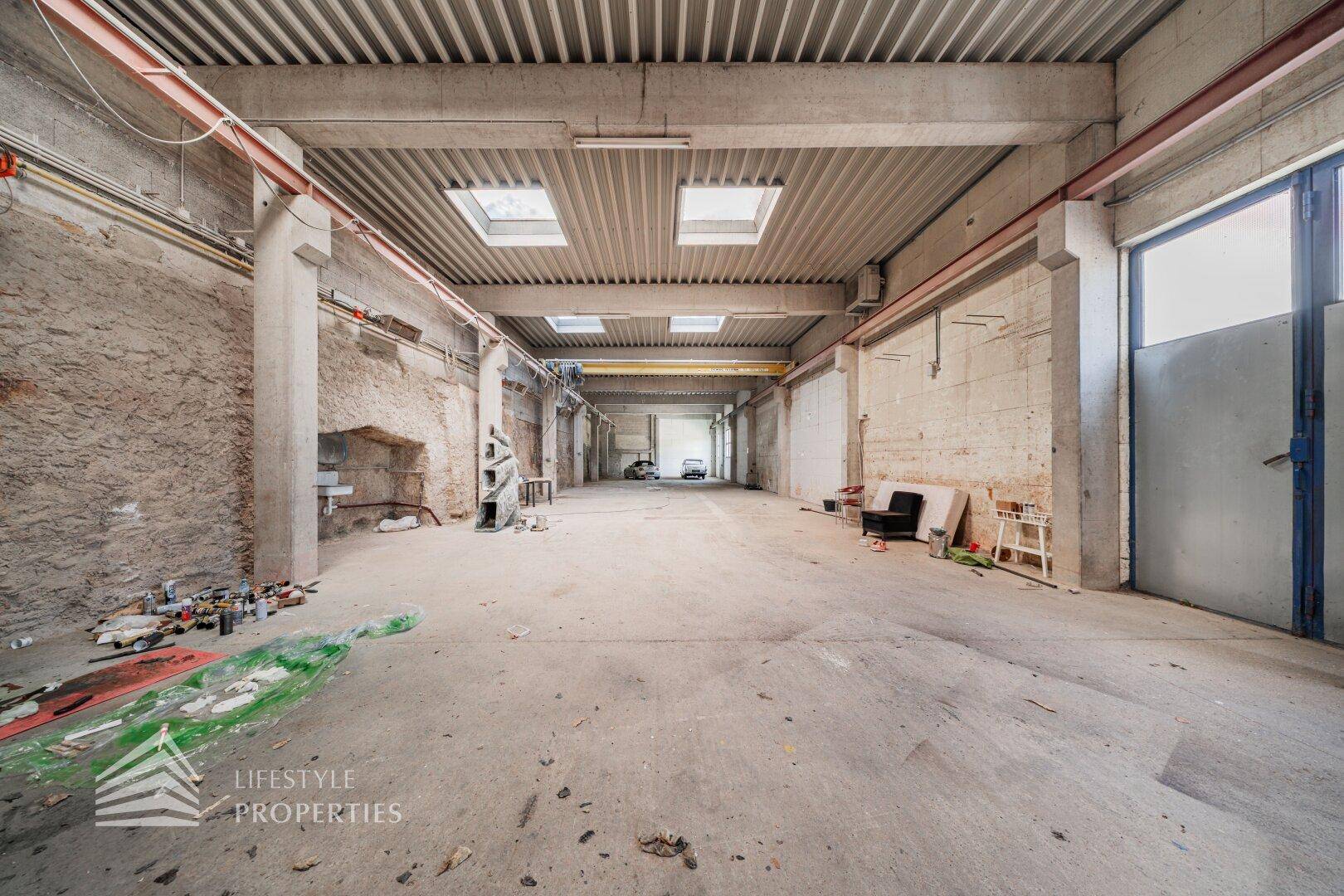 Große, leere Lagerhalle mit Betonboden, hohen Decken und Tageslicht durch Oberlichter.