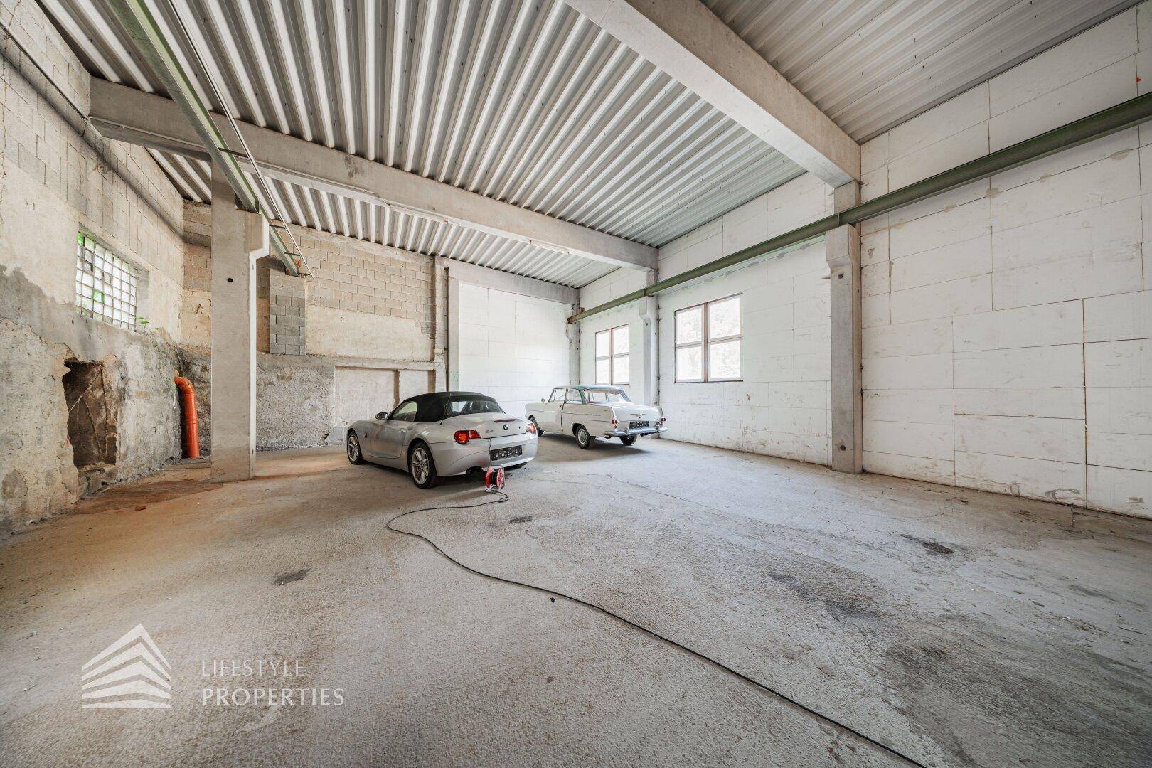 Heller Bereich einer Lagerhalle mit Betonboden und Fenstern, in dem zwei Oldtimer geparkt sind.
