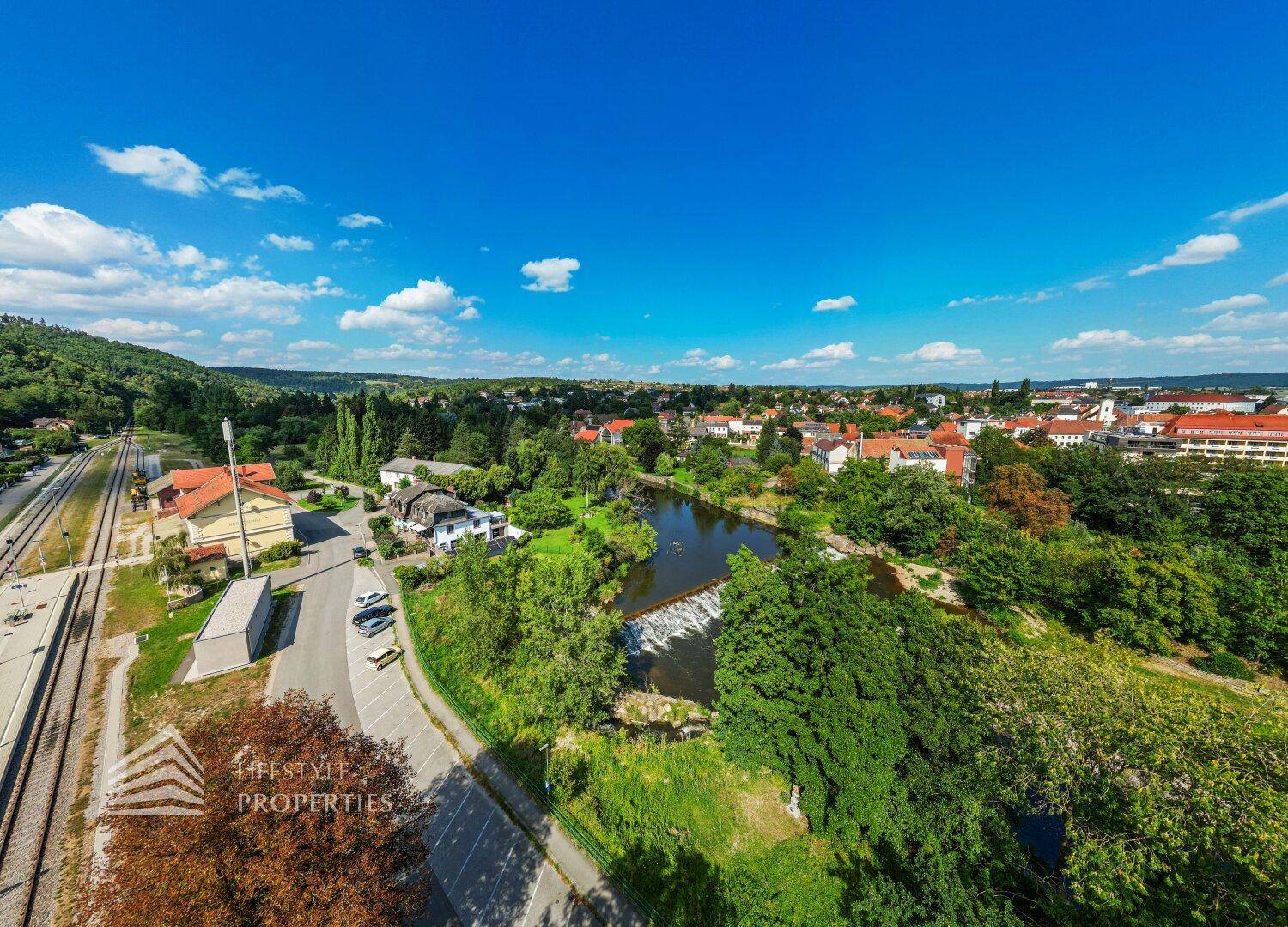 Weitwinkel-Luftaufnahme einer Stadtlandschaft mit Fluss, Bahnlinie und grünen Hügeln im Hintergrund.