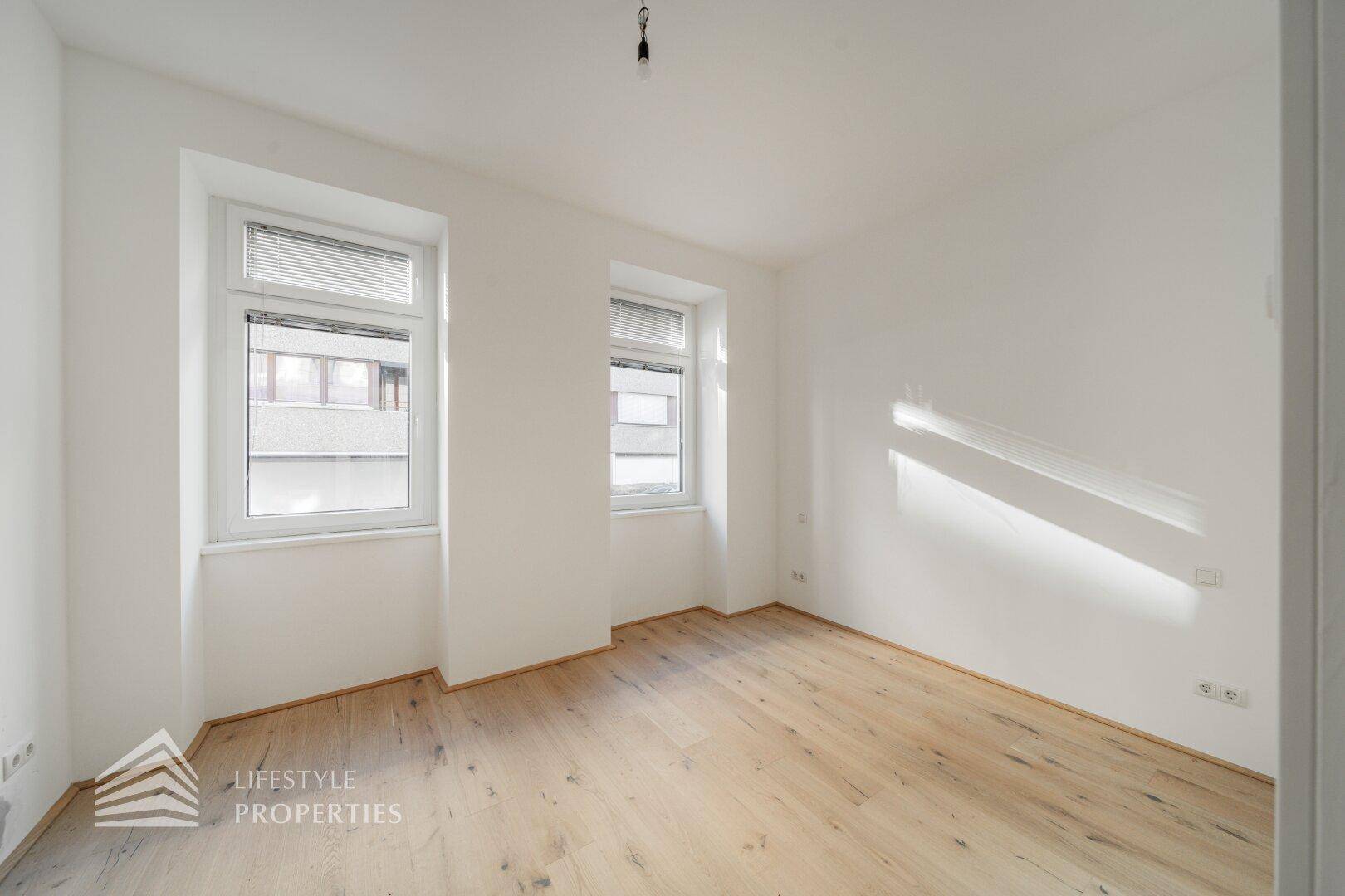 Heller, leerer Raum mit Holzboden und zwei Fenstern, die für natürliche Belichtung sorgen.
