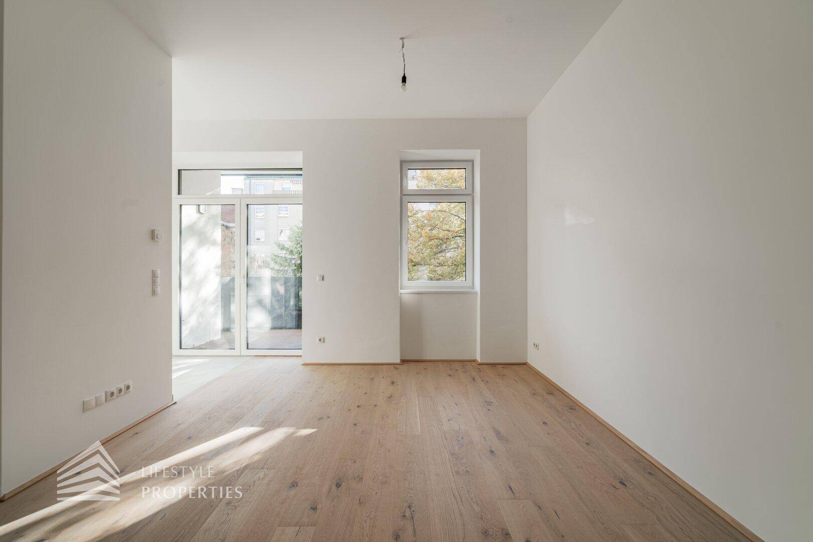 Heller Raum mit Holzboden, einem Fenster und einer Glastür, die Zugang nach draußen bietet.