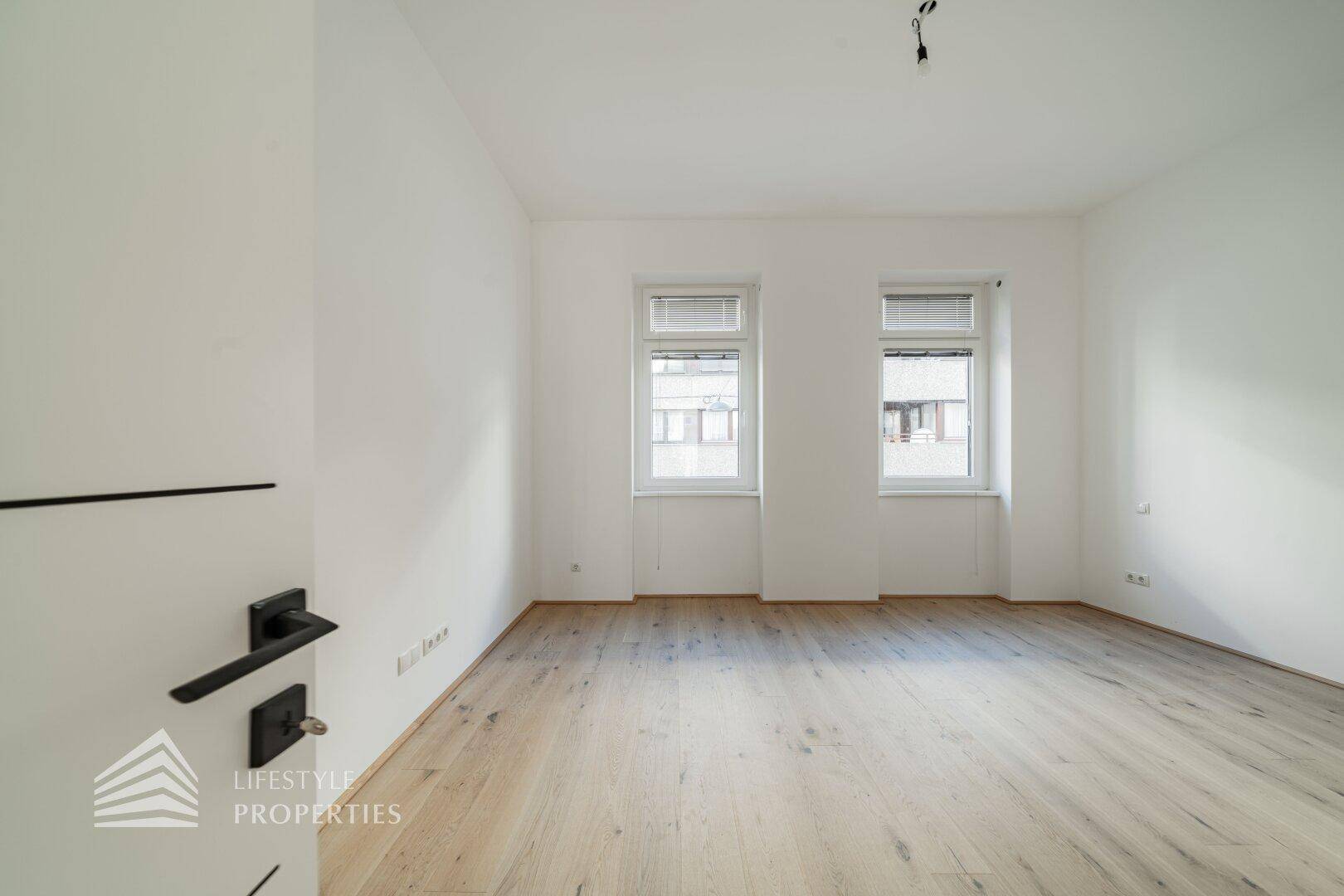 Heller Raum mit Holzboden und zwei Fenstern, die für eine angenehme Atmosphäre sorgen.