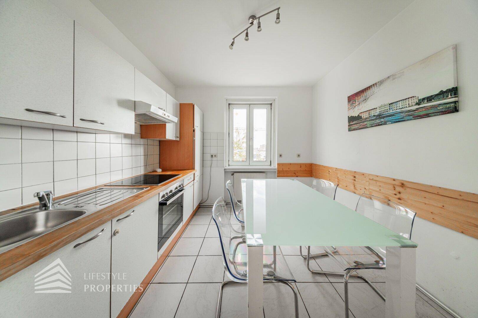 Funktionale Küche mit weißen Schränken, Holz-Arbeitsplatte, Essbereich und Fenster für Tageslicht.
