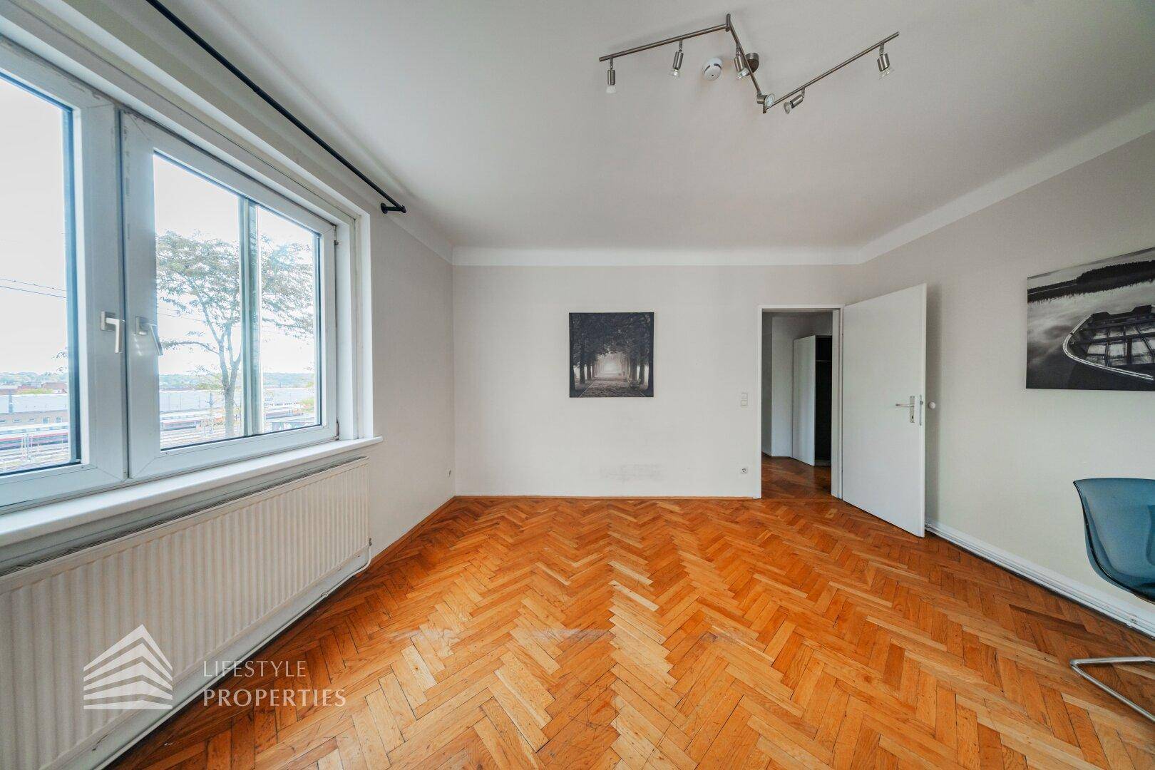 Lichtdurchfluteter Raum mit Fischgrätparkett, großem Fenster und offenem Zugang zu weiteren Zimmern.