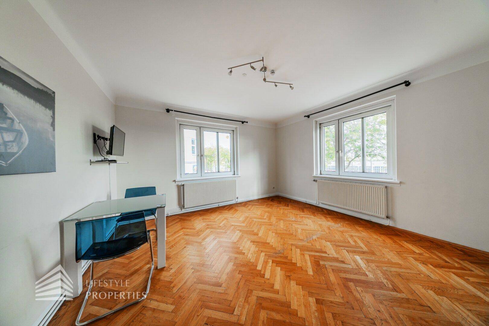 Geräumiges Zimmer mit klassischem Fischgrätparkett und viel Tageslicht durch zwei große Fenster.