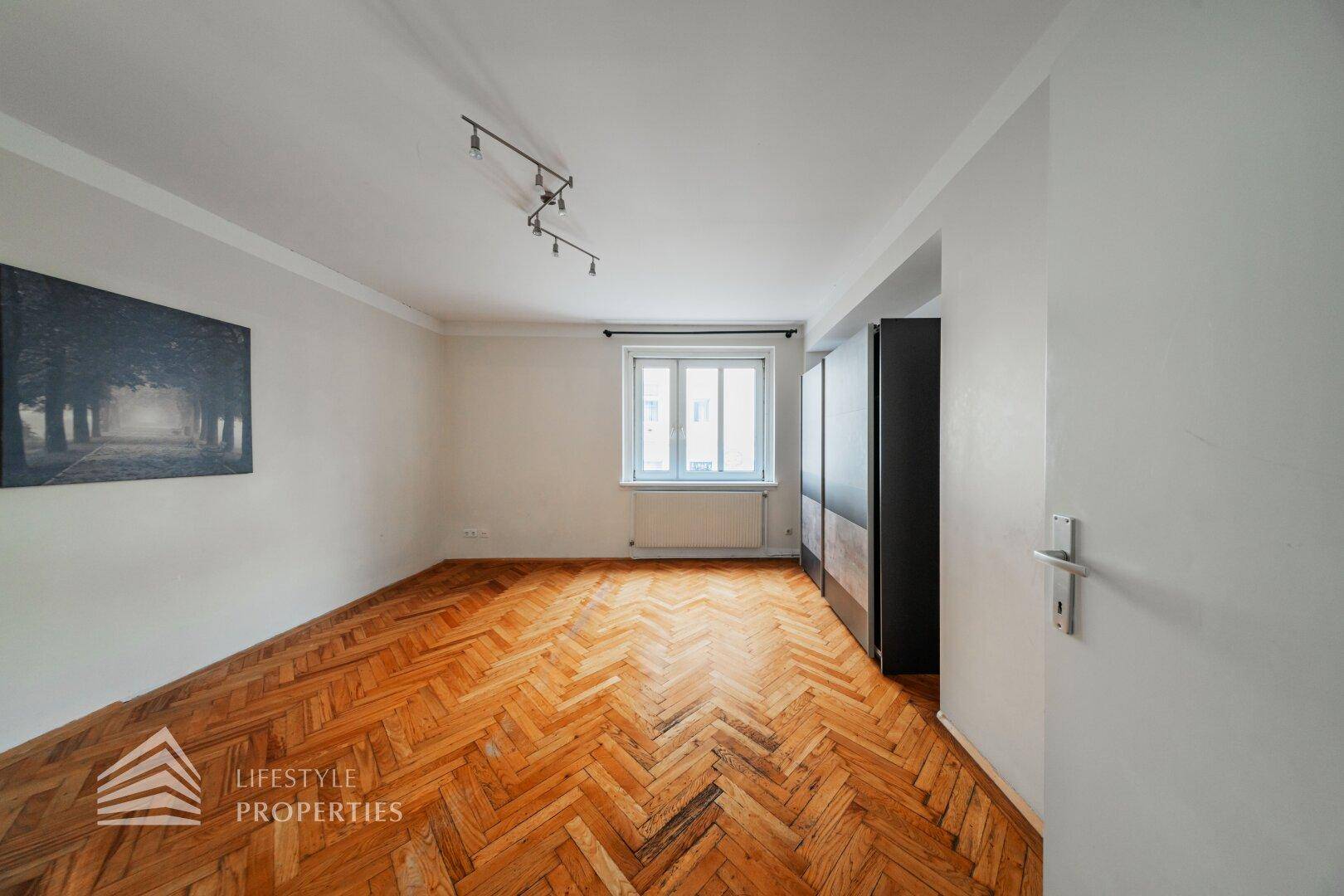 Leeres Zimmer mit Fischgrätparkett, großem Fenster und einem dunklen, freistehenden Kleiderschrank.