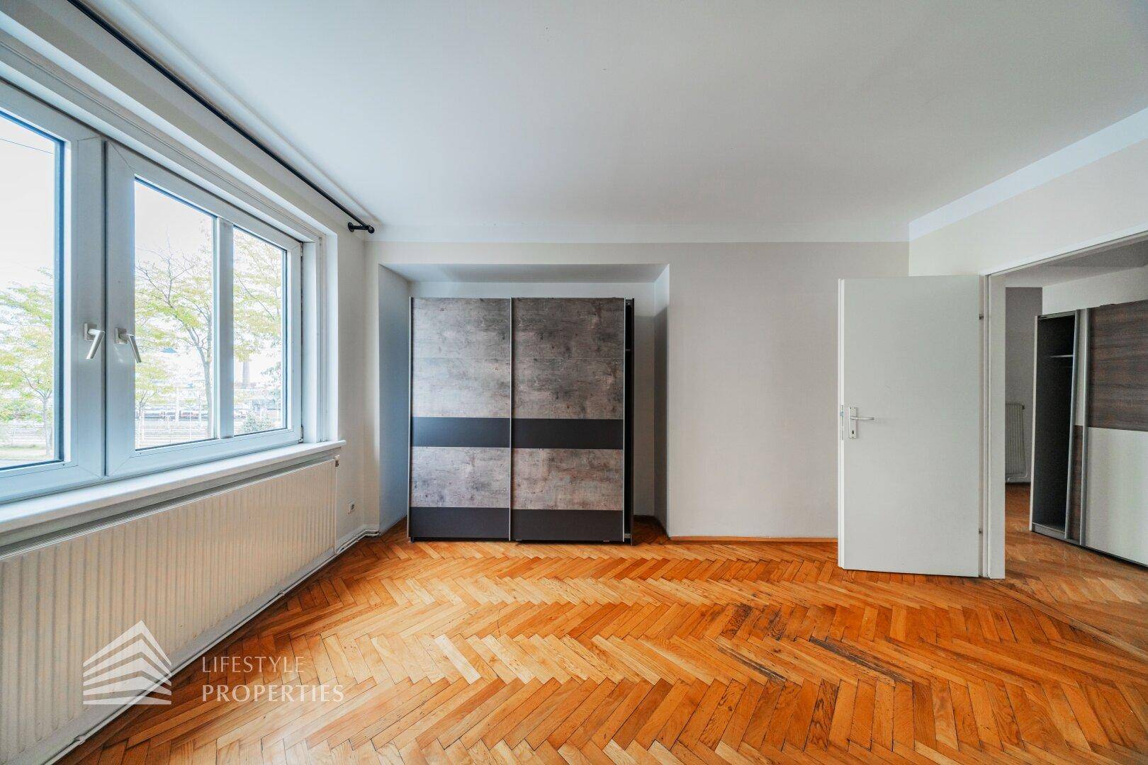 Helles Zimmer mit Fischgrätparkett, großem Fenster und einem modernen, freistehenden Kleiderschrank.