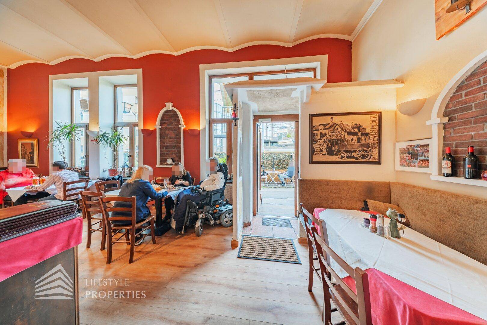 Belebter Restaurantbereich mit Gästen, Holzboden und Zugang zu einem Außenbereich, hell und einladend.