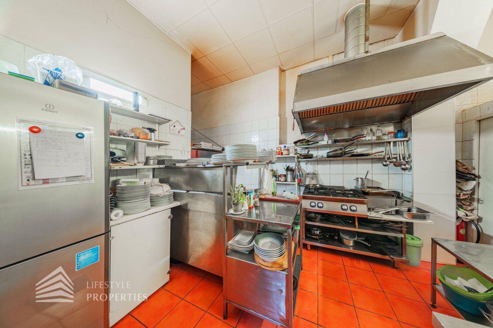 Professionelle Restaurantküche mit großem Dunstabzug, Gasherd, Kühlschrank und orangefarbenen Fliesenboden.