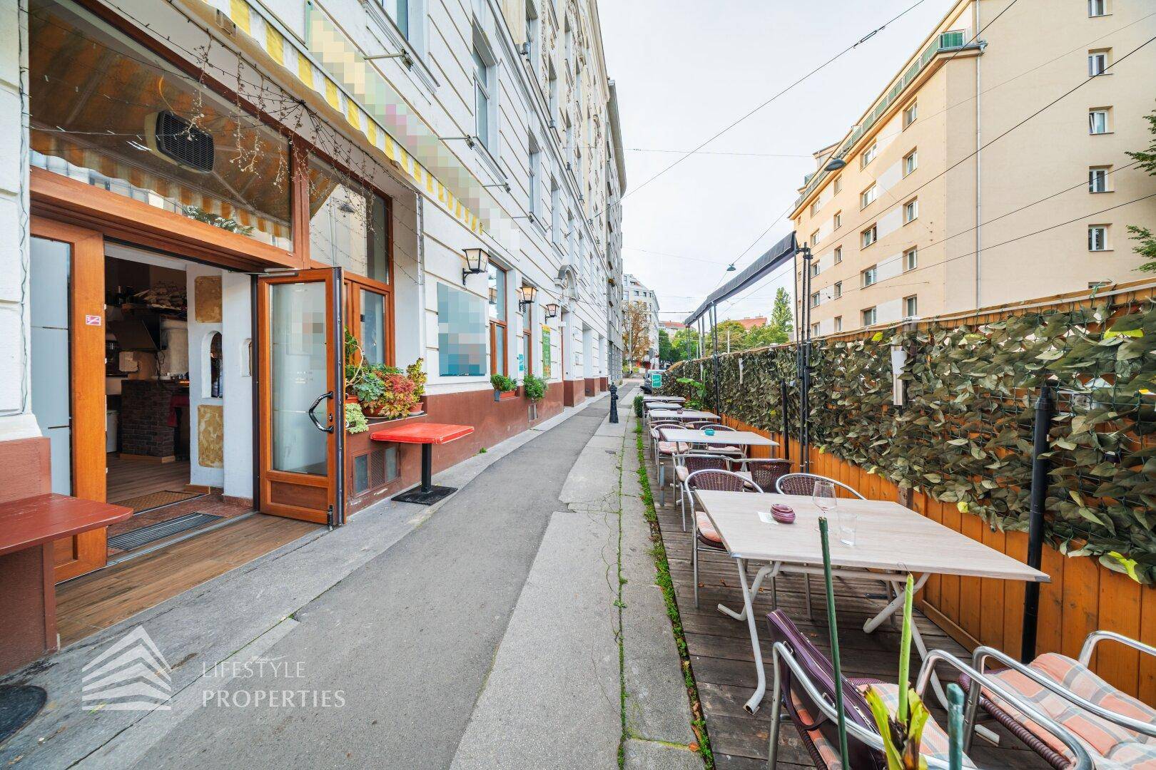 Außenbereich eines Restaurants mit Tischen und Stühlen entlang des Bürgersteigs, umgeben von Gebäuden.