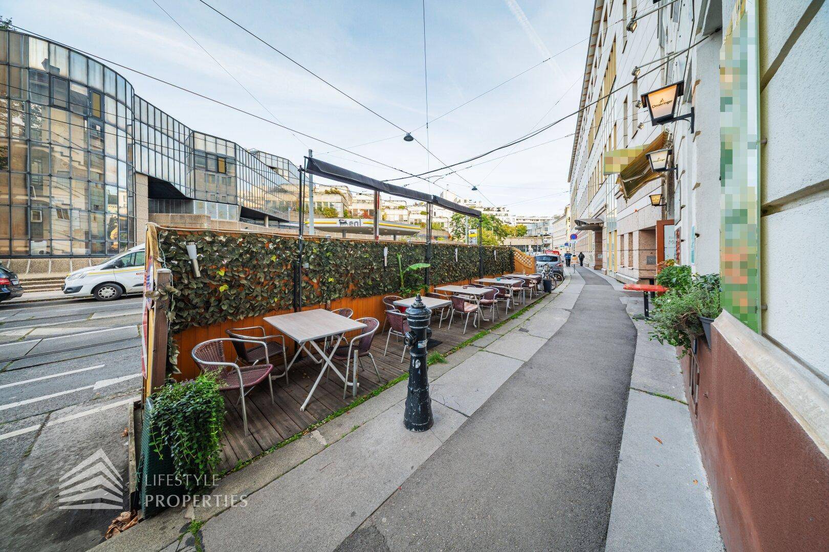 Terrassenbereich eines Restaurants mit Tischen und Stühlen, Blick auf die Straße und umliegende Gebäude.