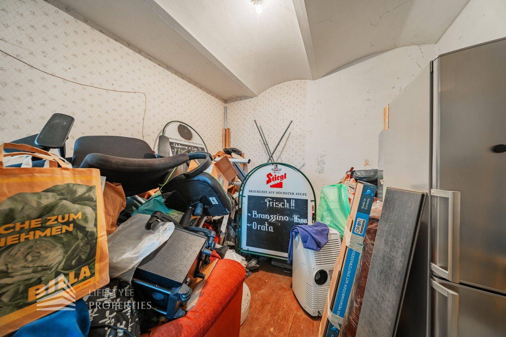 Unaufgeräumter Abstellraum mit verschiedenen Gegenständen, Kühlschrank und schrägen Wänden.