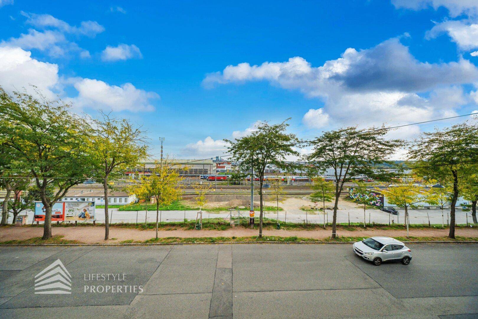 Blick auf eine Straße mit Bäumen, dahinter Bahngleise und städtische Gebäude unter blauem Himmel.