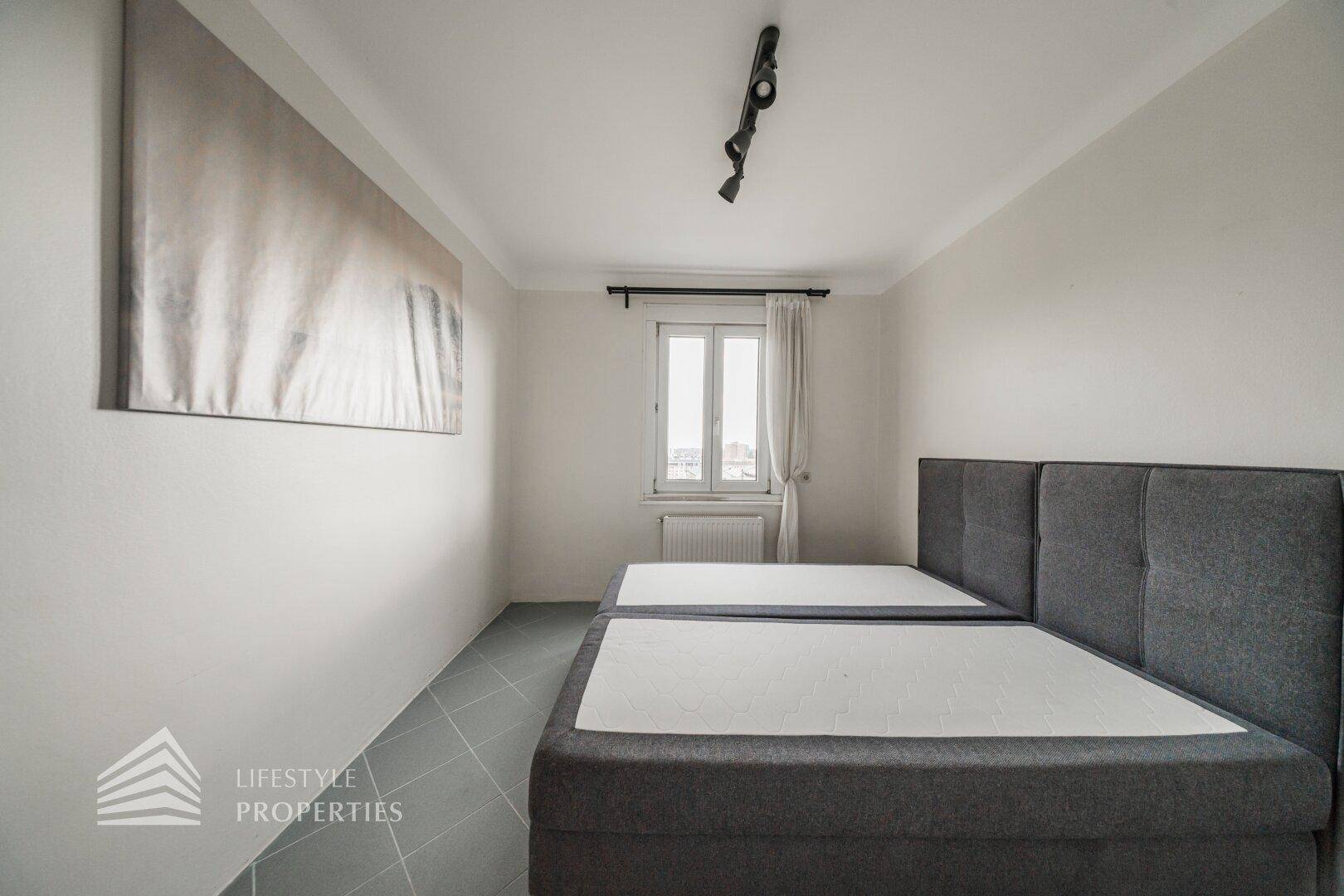 Schlafzimmer mit zwei Einzelbetten, grauem Fliesenboden und einem Fenster für natürliche Belichtung.