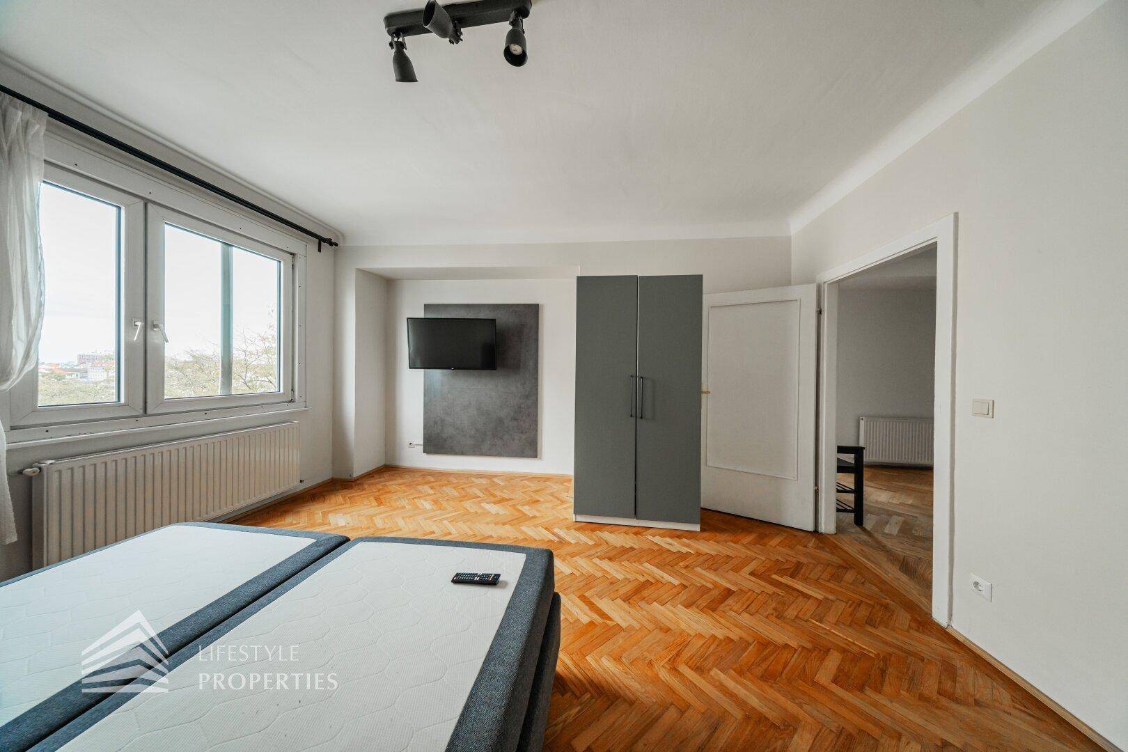 Schlafzimmer mit Fischgrätparkett, Doppelbett, Kleiderschrank und Blick aus dem Fenster.