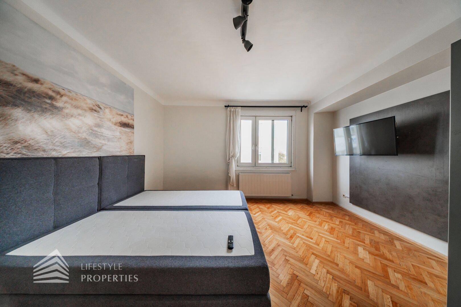 Schlafzimmer mit Doppelbett, Fischgrätparkett und einem Fenster, das Tageslicht hereinlässt.