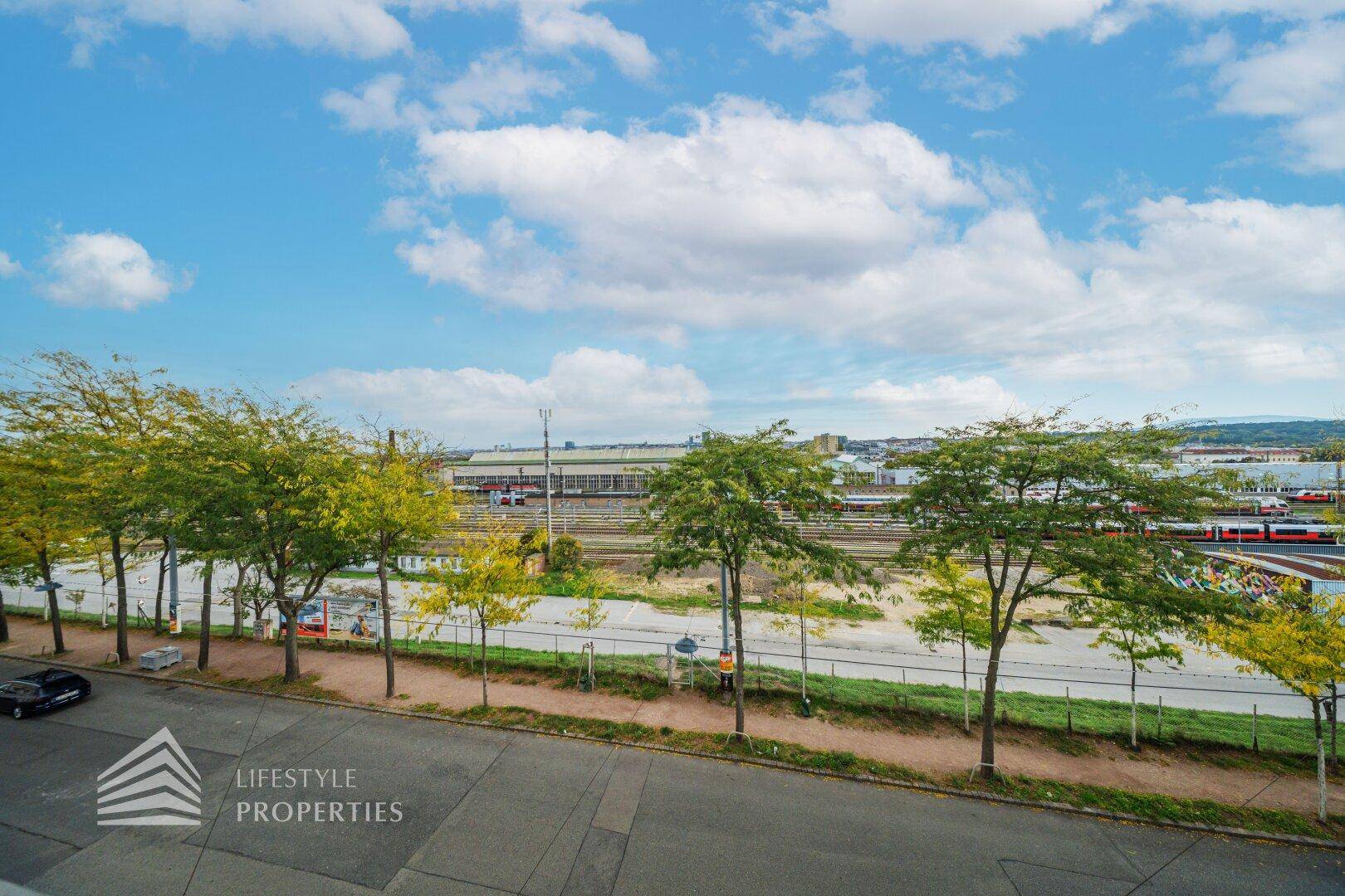 Ausblick auf Bahngleise und Bäume unter blauem Himmel mit Wolken.