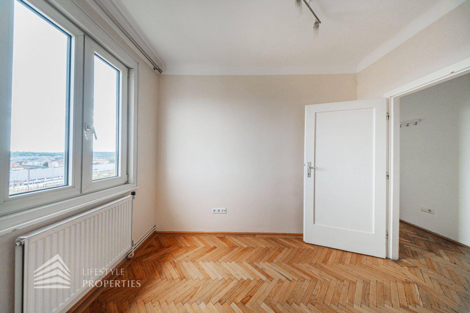 Leeres Zimmer mit großen Fenstern, Fischgrätparkett und viel Tageslicht.