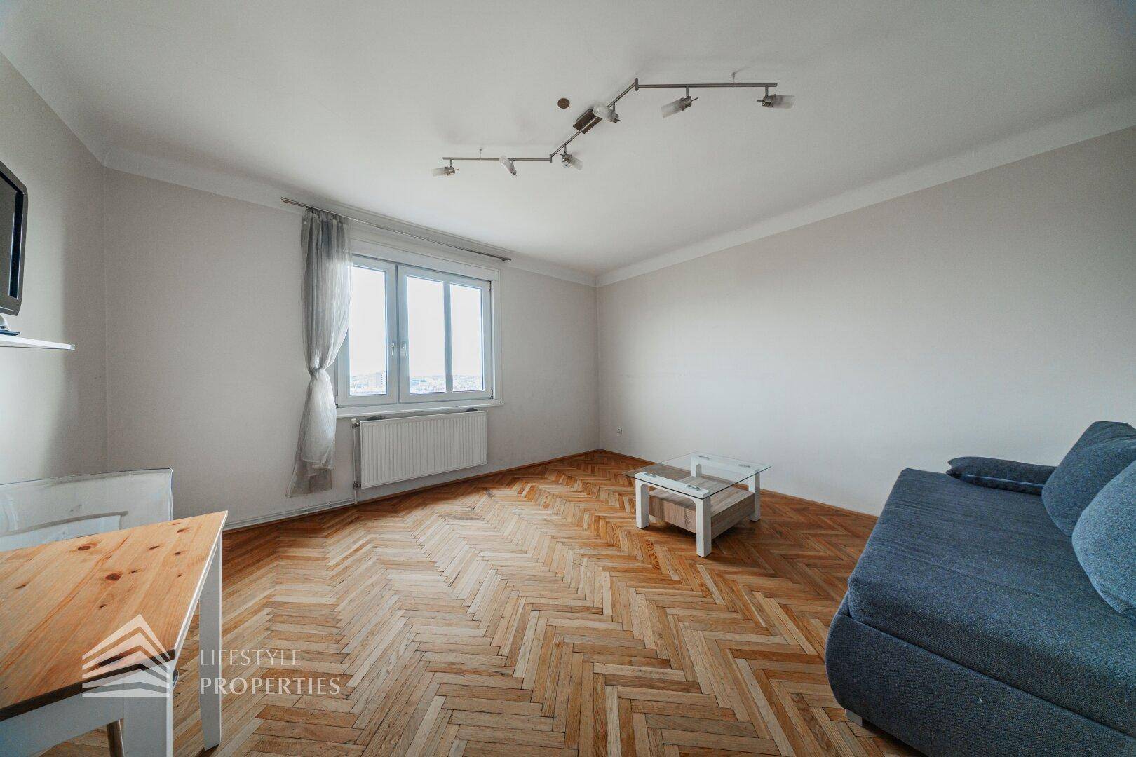 Helles Wohnzimmer mit Fischgrätparkett, bequemer Couch und großem Fenster.