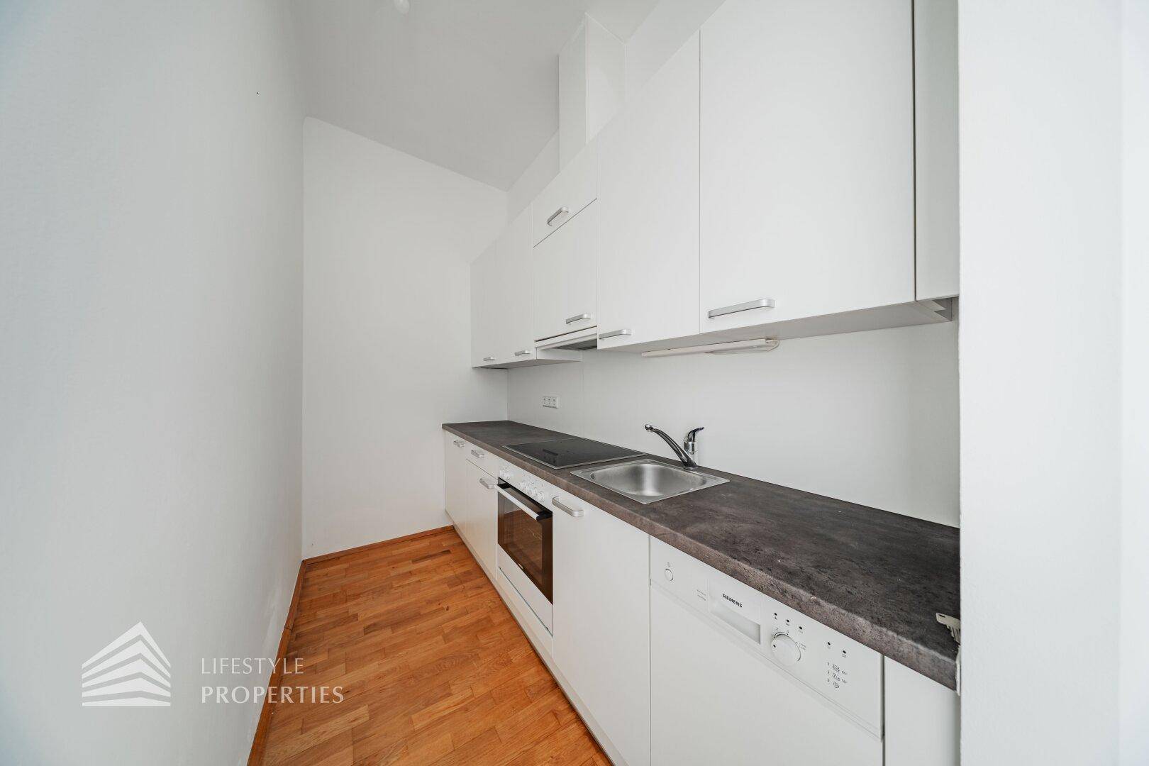 Moderne, schmale Küche mit weißen Schränken, dunkler Arbeitsplatte und Holzboden.