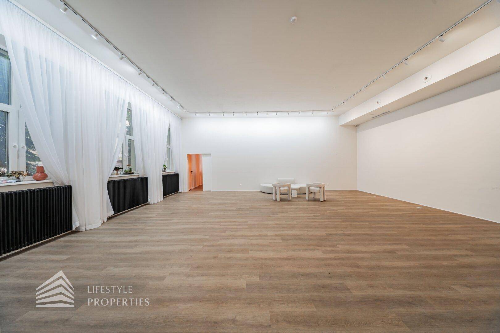 Weitläufiger Mehrzweckraum mit großen Fenstern, eleganten Vorhängen und Holzboden für diverse Aktivitäten.